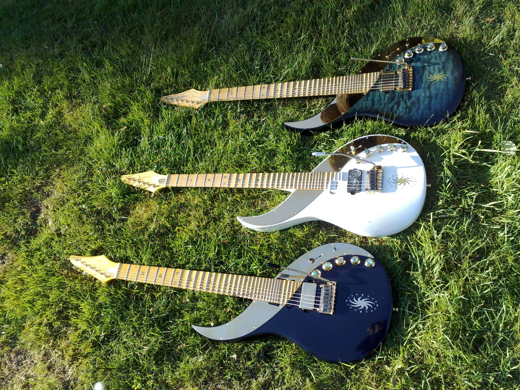 Tres Sky Guitars tumbadas en la hierba: una blanca, una azul acolchada y una azul oscura