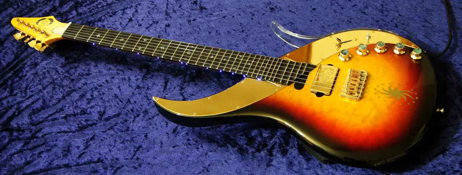 Ebony Sky sunburst en diagonal sobre terciopelo azul, con golpeador dorado y diapasón de ébano